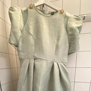 Ljusgrön kortklänning med puffärm och mönster - Superfin ljusgrön klänning från shein med puffiga korta ärmar och subtilt broderat mönster. Klänningen har dekorativa guldknappar vid axlarna och en rund halsringning. Perfekt för dig som gillar en feminin och elegant stil. Använd 1 tillfällen på 2timmar. Storlek M