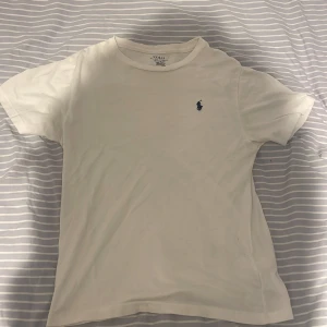 Vit t-shirt från Polo Ralph Lauren - Vit t-shirt från Polo Ralph Lauren med klassisk passform och korta ärmar. Diskret blå logga broderad på bröstet. Tillverkad i mjuk bomull som känns skön mot huden. Perfekt basic-plagg med stilren look. Storlek 150cm.