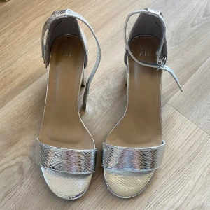 Silvriga klackade sandaler från din sko - Säljer ett par silvriga sandaler med blockklack och öppen tå från xit. Skorna har ett glittrigt, ormskinnsmönstrat material och justerbart spänne runt vristen. Perfekta för att lyfta din outfit med lite extra bling.