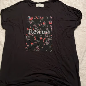 Svart t-shirt med tryck från ADPT - Svart t-shirt från ADPT med coolt tryck framtill där det står 'Rêveuse' och små blommor i rött, rosa och blått. T-shirten har korta ärmar och en avslappnad passform. Perfekt för dig som gillar en chill och trendig stil.