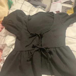 Svart blus med puffärm från Gina Tricot - Supersöt svart blus från Gina Tricot i ribbad bomullsmix. Modellen har korta puffärmar, markerad midja och två knytband framtill. Urringad hals och volang nedtill ger en romantisk vibe. Perfekt att styla med jeans eller kjol för en trendig look.