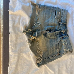 Ljusblå jeansshorts med fransar - Säljer ett par ljusblå jeansshorts med råa, fransiga kanter och slitna detaljer. Klassisk femficksmodell med knapp och dragkedja framtill. Perfekta för sommaren och ger en cool, avslappnad look.