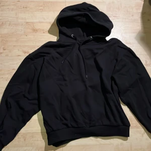 Svart hoodie - Säljer en klassisk svart hoodie med huva och dragsko. Tröjan har långa ärmar och bred mudd vid ärmslut och nederkant. Perfekt för en avslappnad stil. Perfekt tunn till sommaren.