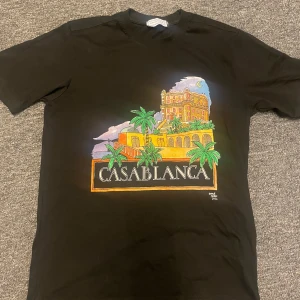 Svart t-shirt med tryck från Casablanca - Säljer en svart t-shirt från Casablanca med färgglatt tryck av byggnad, palmer och texten 'Casablanca' på framsidan. Klassisk passform och rund hals. Perfekt för dig som vill sticka ut med en snygg grafisk tee.