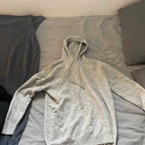 Ljusgrå hoodie i ullmix - Mysig ljusgrå hoodie med huva och snörning. Tröjan är stickad i en mjuk ullmix och har långa ärmar samt ribbade muddar vid ärmslut och nederkant. Perfekt för kyliga dagar och enkel att matcha med jeans eller mjukisbyxor.
