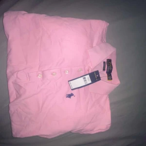 Rosa pikétröja från Polo Ralph Lauren - Snygg rosa pikétröja från Polo Ralph Lauren i klassisk modell med krage och knappar framtill. Tröjan har mörkblå broderad logga på bröstet och är gjord i mjuk bomull. Perfekt för en fräsch och sportig look.