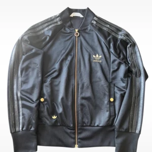 Svart Adidas track jacket med gulddetaljer - Säljer en svart Adidas track jacket med guldfärgade detaljer och dragkedja. Jackan har klassiska tre ränder längs ärmarna, ribbade muddar och Adidas logga både fram och bak. Perfekt för en sportig och stilren look.