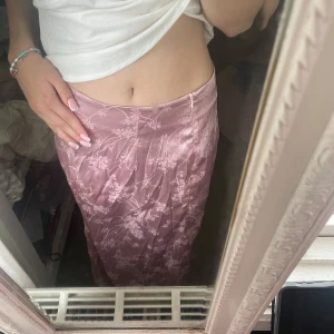 Ljusrosa blommig satinkjol - Supersnygg långkjol i ljusrosa satin med blommigt mönster. Kjolen är i storlek M men passar mer som S. Perfekt för dig som vill ha en romantisk och trendig look.