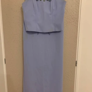 Ljusblå långklänning med snörning - Supersöt ljusblå långklänning i tvådelad look med smala axelband och snörning i ryggen. Toppen har små broderade blommor framtill och kjolen är rak med slits bak. Perfekt för dig som vill sticka ut med en romantisk vibe. Passar small och liten medium. Finns liten ljus fläck på kjolen. Går säkert att tvätta bort. 