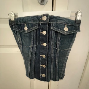 Mörk jeans topp  - Snygg mörkblå jeanstopp med fransig kant och coola silverfärgade knappar framtill. Kjolen har två fickor med lock och markerade sömmar som ger en trendig look. Perfekt för dig som gillar denim och vill sticka ut lite extra.
