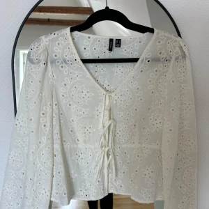 Vit blus med snörning från vero Moda - Superfin vit blus från vero Moda med broderade detaljer och v-ringning. Blusen har snörning framtill och långa ärmar. Tunt bomullstyg med broderade mönster ger en luftig och romantisk känsla. Perfekt för vår och sommar!