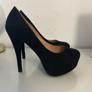 Svarta pumps med platå från Tulipano - Svarta pumps med hög klack och platåsula från Tulipano i storlek 37. Skorna har rund tå och är klädda i mockaliknande material. I det stora hela är skorna i bra skick men klackarna är något slitna vilket syns på bilderna. 