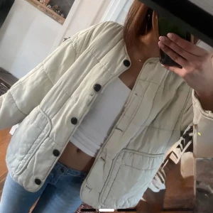 Vit quiltad jacka med svarta knappar - Säljer en vit quiltad jacka med svarta knappar och rund halsringning. Jackan har en boxig passform och stora fickor framtill. Perfekt att slänga över en t-shirt för en avslappnad look.