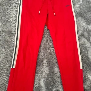Röda mjukisbyxor från Hugo Boss - Snygga röda mjukisbyxor med vita och svarta revärer längs sidorna och snörning i midjan. Perfekta för en sportig look eller chill hemma. Liten BOSS-logga på ena höften.