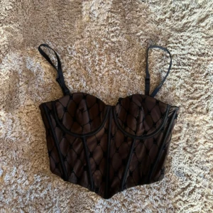 Svart korsettopp från Amisu - Säljer en svart korsettopp från Amisu med transparent mesh och broderade detaljer. Toppen har justerbara axelband och dragkedja i sidan. Perfekt för att lyfta vilken outfit som helst!