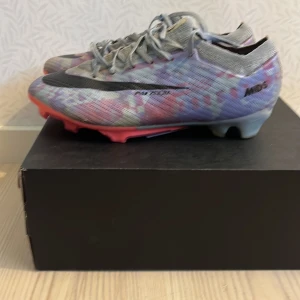 Nike Mercurial fotbollsskor i grått och rosa - Säljer ett par Nike Mercurial fotbollsskor med grå bas och inslag av rosa och lila. Skorna har snörning, svart Nike-logga och platt sula med dobbar för gräsplan. Perfekta för dig som vill sticka ut på planen! Skorna är lite trasiga på innersidorna vid Nike märket 