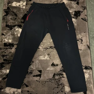 Svarta mjukisbyxor Race Marine M - Svarta mjukisbyxor från Race Marine med elastisk midja och snörning. Snygga röda detaljer vid fickorna och tryckt text på ena benet. Sköna och stilrena byxor i mjukt material, perfekta för chill eller träning.