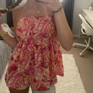 Paisleymönstrad rosa tubtopp - Säljer en färgglad tubtopp med paisleymönster i rosa, orange och vitt. Toppen är bandeau-modell och har en luftig, flowy passform som är perfekt för varma dagar. Resår upptill för att sitta på plats. Superfin till shorts eller kjol!