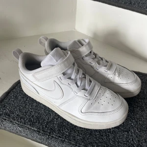 Vita Nike sneakers med kardborre - Säljer ett par helvita Nike sneakers med kardborreband och snörning. Skorna har perforerade detaljer på tån för ventilation och en klassisk Nike-logga på sidan. Perfekta för en clean och sportig look. Storlek 34 Innermått 21,5 cm