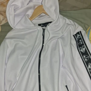 Moose knuckles hoodie med svarta detaljer och mönstrade band - Vit hoodie med dragkedja och svarta detaljer. Svarta mönstrade band längs ärmarna och svarta ribbade muddar vid ärmslut och nederkant. Huva och fickor med dragkedja.