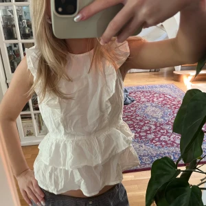 Vit volangblus med knyt i ryggen - Säljer en söt vit blus med volangdetaljer och ärmlös design. Blusen har ett fint knyt i ryggen och dubbla volanger vid midjan för en romantisk look. Perfekt till sommaren!