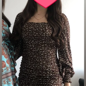 Brun blommig kortklänning med långa ärmar - Supersöt brun kortklänning med små vita blommor, smockad överdel och långa puffärmar. Fyrkantig ringning och figurnära passform ger en trendig look. Perfekt för dig som gillar romantiska detaljer och vill sticka ut.