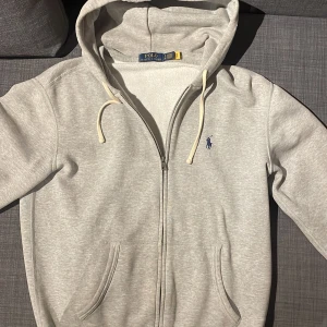 Grå hoodie från Polo Ralph Lauren - Snygg ljusgrå hoodie från Polo Ralph Lauren med dragkedja, huva och vita snören. Klassisk broderad logga i mörkblått på bröstet och två fickor framtill. Perfekt för chill dagar och enkel att matcha med jeans eller joggers.