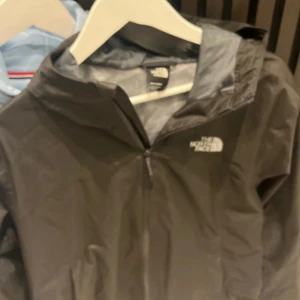 Svart vindjacka från The North Face - Svart vindjacka från The North Face i storlek M. Jackan har huva, dragkedja framtill och meshfoder på insidan. Perfekt för blåsiga dagar och enkel att slänga på sig när vädret svänger. Klassisk logga på bröstet och stilren design.