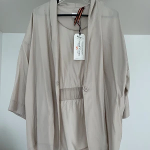 Beige kimono med knyt från Lindex - Säljer en beige kimono från Lindex med avslappnad passform och trekvartsärm. Kimonon har en enkel knäppning framtill och en lös, öppen design med knytskärp i midjan. Perfekt för lager-på-lager och stilrena outfits.