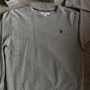 Grå polo tröja - Snygg grå långärmad tröja från polo med klassisk rund hals och broderad logga på bröstet. Tillverkad i mjuk bomullsmix med 10% viskos för extra komfort. Perfekt för en clean och stilren look.