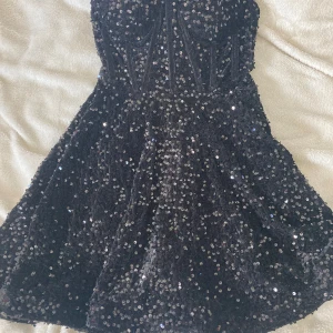 Svart paljettklänning axelbandslös - Glittrig svart kortklänning med paljetter över hela tyget. Axelbandslös modell med markerad midja och lätt utställd kjol. Perfekt för dig som vill sticka ut och glänsa på festen.