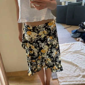 Blommig svart midikjol  - Supersöt svart midikjol med blommigt mönster i vitt och gult från Ü. Kjolen har volangkant nertill och en flowy passform som ger en skön och avslappnad vibe. Perfekt för dig som gillar färg och mönster💗