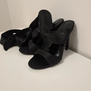 Svarta glittriga klackar med band 12cm  - Snygga svarta klackar med öppen tå och breda band täckta av små svarta stenar som ger en glittrig effekt. Skorna har höga klackar och långa band att knyta runt vristen. Perfekta för dig som vill sticka ut med en elegant och trendig look.