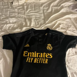 Real Madrid svart matchtröja Adidas - Snygg svart Real Madrid fotbollströja från Adidas med guldiga detaljer och klubbmärke på bröstet. Klassisk passform med korta ärmar och tre ränder på axlarna. Tillverkad i lätt och ventilerande material, perfekt för match eller träning.