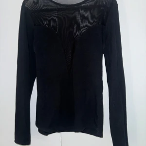 Svart långärmad topp med meshdetalj - Snygg svart långärmad topp med transparent mesh upptill och över axlarna. Rund halsringning och figurnära passform. Perfekt för dig som vill ha en stilren men ändå lite edgy look. Materialet känns mjukt och stretchigt. Storlek M.