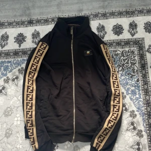 Svart kofta från Fendi - Svart kofta  från Fendi med ikoniska FF-loggor i beige längs ärmarna och dragkedja framtill. Koftan har hög krage, ribbade muddar och coolt Fendi-märke på bröstet. Tillverkad i 100% bomull för en skön känsla och snygg passform. 