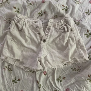 Vita jeansshorts med knappar från Zara - Snygga vita jeansshorts från Zara med hög midja, råa kanter och flera knappar framtill. Shortsen har fickor både fram och bak samt en cool, lite sliten look. Perfekta för sommaren och lätta att matcha med allt.Köpta i New York 