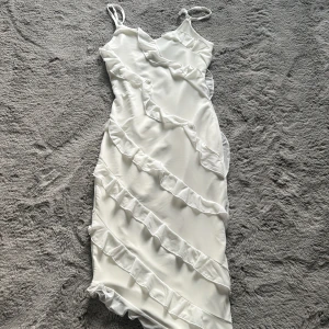 Vit långklänning med volangdetaljer - Superfin vit långklänning med smala axelband och volanger som går diagonalt över hela klänningen. Snyggt figurnära passform och tillverkad i ett glansigt, mjukt tyg. Har använts en gång!☺️