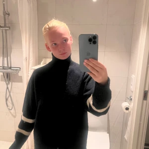 Svart stickad polotröja med ränder - Svart stickad polotröja med breda vita ränder på ärmarna. Tröjan har hög hals och en loose passform, perfekt för kyliga dagar. Ärmarna är långa och muddade vid handlederna. Enkel men stilren design som funkar till många outfits.nypris 449