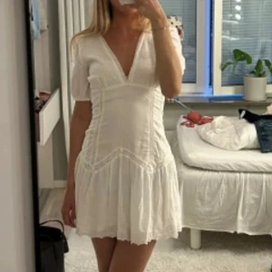 Vit kort sommarklänning från Zara - Superfin vit kort klänning från Zara med v-ringning, broderade detaljer och lätt puffärm. Klänningen har volangkant nertill och är tillverkad i bomull, perfekt för varma dagar. Söt och luftig modell med romantisk känsla.