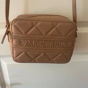 Beige quiltad axelväska från Valentino - Säljer en snygg beige axelväska från Valentino med quiltad design och tydlig logga framtill. Väskan har justerbar axelrem, dragkedja i guld och rymligt fack inuti. Perfekt för dig som vill ha en stilren och trendig väska till vardags.
