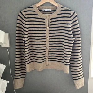 Randig kofta från Zara i beige och svart - Superfin kofta från Zara i beige med svarta ränder. Den har rund hals, guldiga knappar framtill och är stickad i ett mjukt material. Perfekt till lager-på-lager och funkar året runt. Långärmad och med ribbade muddar för extra mysfaktor.