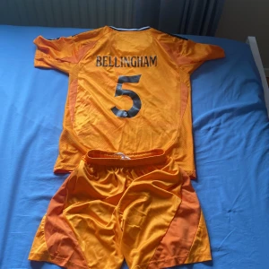 Real Madrid Bellingham matchställ orange - Säljer ett Real Madrid matchställ med Bellingham och nummer 5. Tröjan är orange med svarta detaljer, kortärmad och har klubbmärke, Adidas-logga och sponsortryck. Shortsen är också orange med svarta tryck och klubbmärke. Materialet är lätt och andas, perfekt för fotboll.