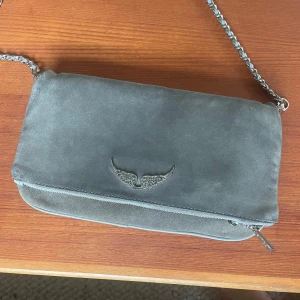 Grå mocka axelväska Zadig & Voltaire - Snygg grå axelväska i mocka från Zadig & Voltaire med silverfärgad kedjerem och dragkedja. Väskan har en ikonisk vingdetalj framtill och praktiskt ytterfack med dragkedja. Perfekt storlek för det viktigaste och stilren look. Köpt 2500 säljer för 1000kr