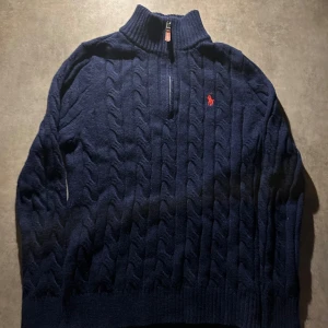 Marinblå kabelstickad tröja Polo Ralph Lauren - Kabelstickad marinblå tröja från Polo Ralph Lauren med halv dragkedja och hög krage. Klassisk röd logga broderad på bröstet. Långärmad och ribbade muddar för extra stilpoäng. Perfekt för lager-på-lager och snygg till jeans. Hör av vid minsta fundering 💫