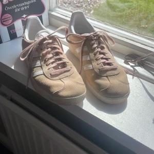 Rosa/beige Adidas Gazelle sneakers strl 36 - Snygga Adidas Gazelle sneakers i beige/rosa mocka med vita detaljer och klassiska tre ränder på sidorna. Skorna har rosa snören och vit sula, samt rund tå. Perfekta för dig som gillar en clean och stilren look med en touch av retro. Skorna är använda få gånger.