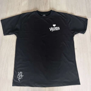Svart Valerra t-shirt med tryck - Svart t-shirt från Valerra med vit logga och hjärta på bröstet samt coolt tryck nere vid fållen. Klassisk rund hals och korta ärmar. Perfekt för dig som gillar streetwear och enkel stil.