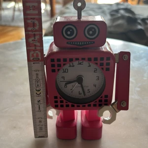 Rosa robotklocka från Moretime - Unik bordsklocka i form av en röd robot från Moretime. Klockan har ett vitt ur med svarta siffror och roboten har coola detaljer som rörliga armar och skruvliknande händer. Perfekt som dekoration eller för att hålla koll på tiden på skrivbordet.