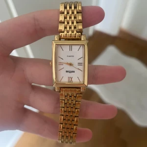 Guldig rektangulär klocka lite stor - Stilren klocka med rektangulärt ur och klassisk länk i guldtonad metall. Urtavlan är vit med romerska siffror och tunna visare. Perfekt accessoar för dig som gillar en tidlös och elegant look.