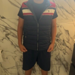 Blå och röd dunväst från U.S. Polo Assn. - Snygg dunväst från U.S. Polo Assn. i mörkblått med röda och vita detaljer. Västen har quiltad design, dragkedja framtill och broderad logga på bröstet. Perfekt för lager-på-lager och har en sportig vibe med sina breda färgblock.